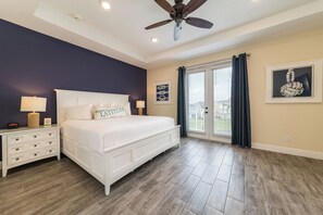 Une chambre avec un lit blanc, des murs bleu foncé, un ventilateur de plafond et une vue sur l’extérieur, visible à travers une porte vitrée.
