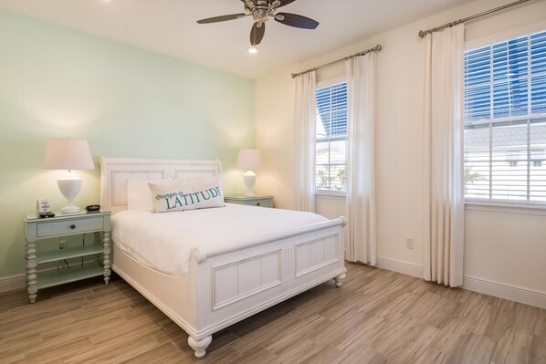 A bedroom with a white bed, two nightstands with lamps, a ceiling fan, and a window with blinds.