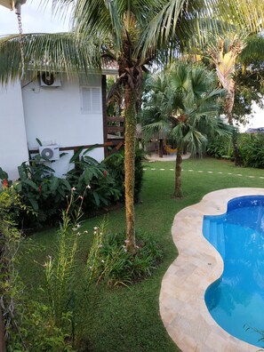 Un patio trasero con piscina, un edificio blanco y una unidad de aire acondicionado, y varias plantas tropicales.