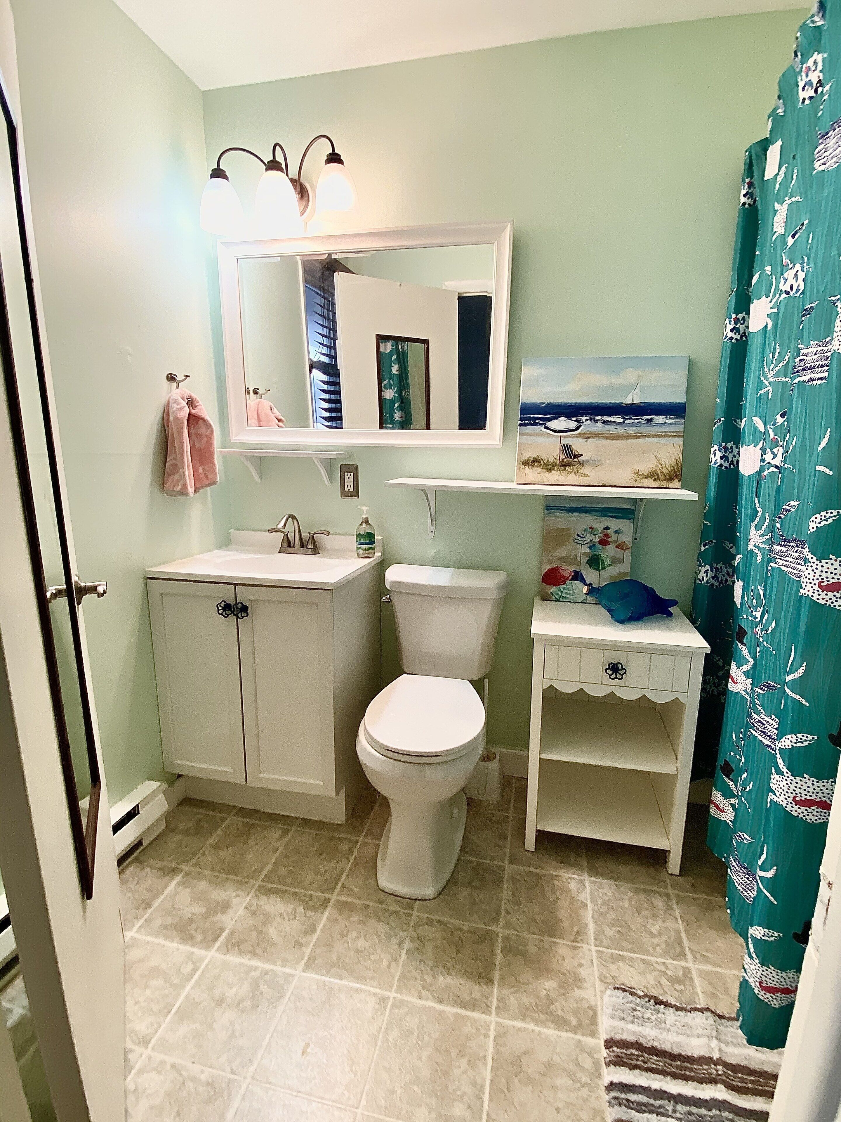A bathroom with a sink, toilet, and a mirror above the sink. There is a towel hanging on the wall and a painting above the mirror.