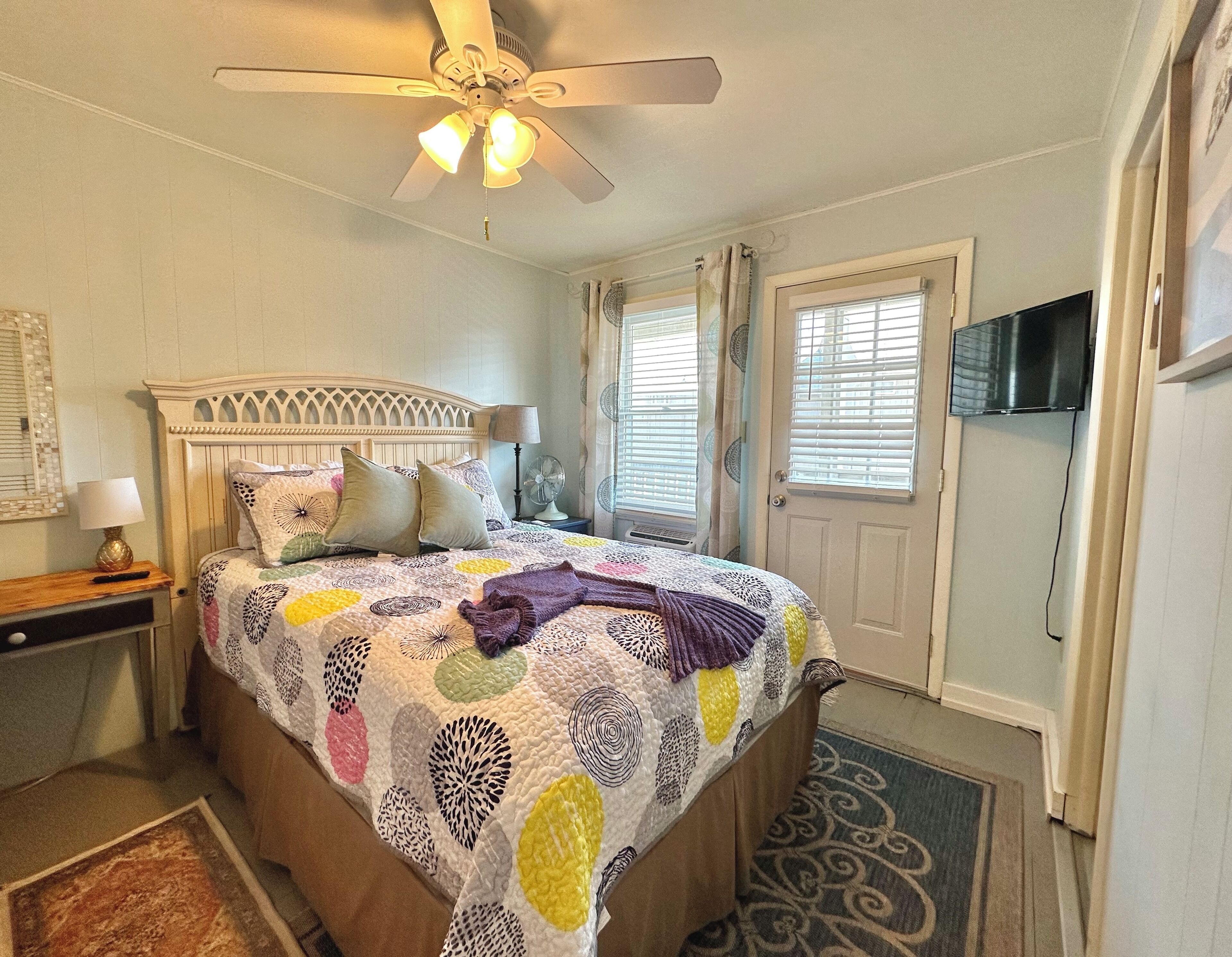 A bedroom with a bed, a ceiling fan, a nightstand, a lamp, a TV, and a window with blinds.