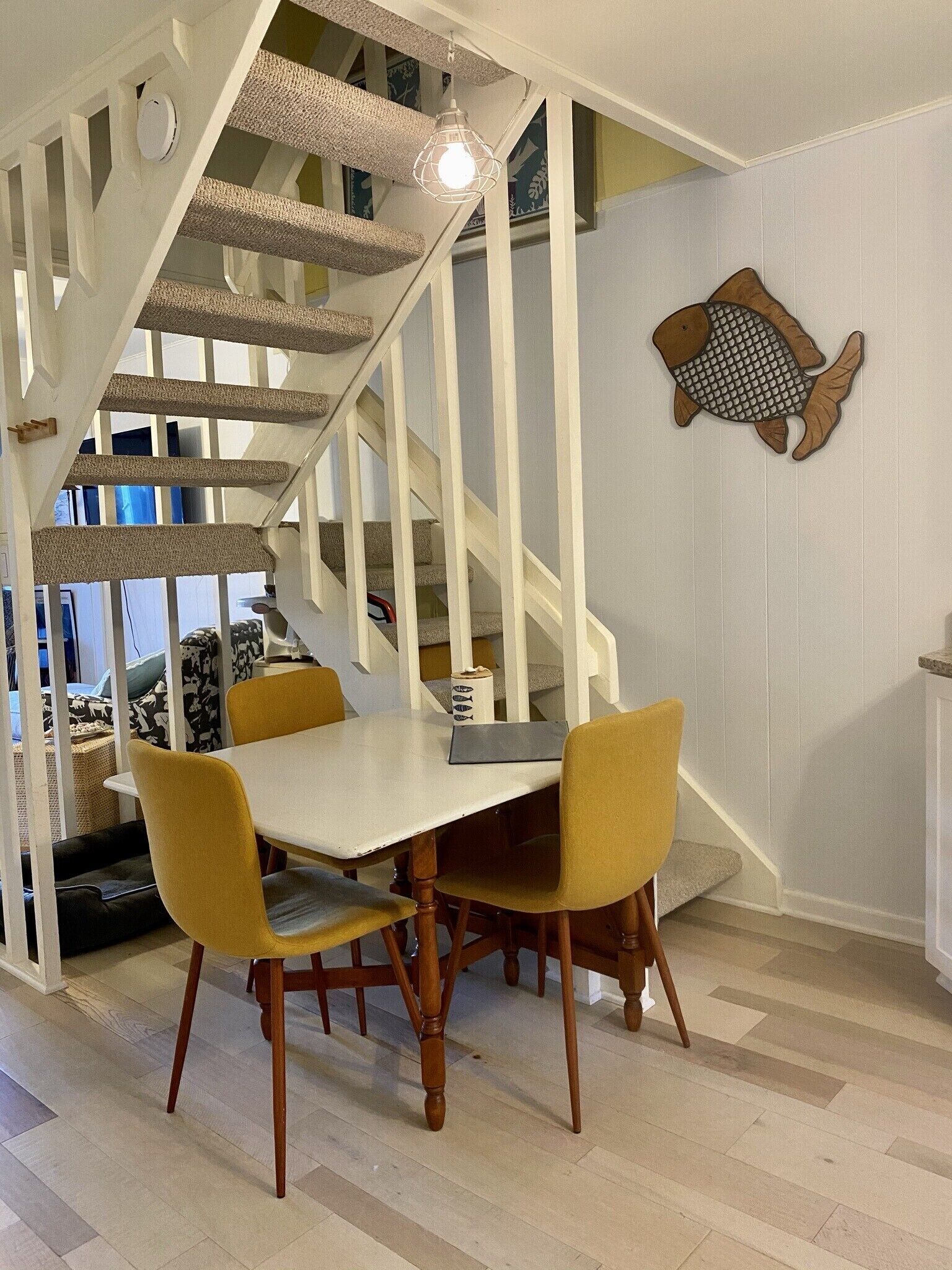 Un coin repas avec une table blanche et des chaises jaunes, un escalier avec des rampes blanches et une décoration murale en forme de poisson.