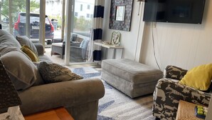 Un salon comprenant un canapé, un pouf et un fauteuil à motifs, un téléviseur à écran plat et une porte vitrée donnant sur l’extérieur.