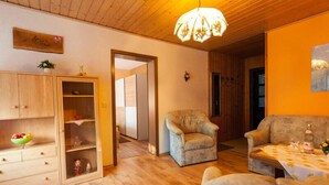 Ein gemütliches Wohnzimmer mit Holzbalkendecke, einer Deckenleuchte, einem Sofa, einem Sessel und einem Esstisch mit Flasche und Gläsern.