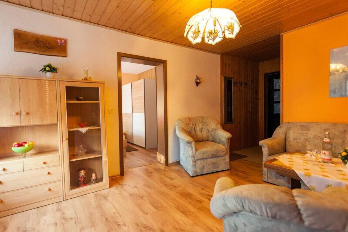 A cozy living room with a wooden ceiling, a chandelier, a sofa, a chair, and a dining table set with a bottle and glasses.