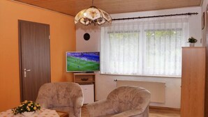 Ein Wohnzimmer mit zwei Sesseln, einem Fernseher, einem Tisch mit Blumenarrangement, einer Holzbalkendecke und einem Fenster mit durchsichtigen Vorhängen.