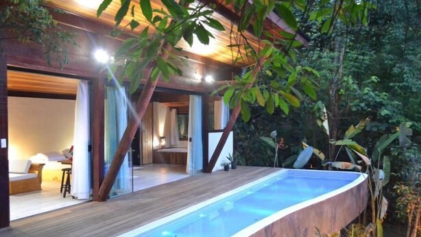 A modern outdoor pool area with a wooden deck and glass walls, surrounded by lush greenery. - Hotel Warabi (Resende)