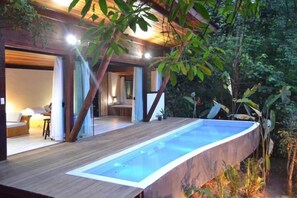 Uma área de piscina moderna ao ar livre com deck de madeira e paredes de vidro, cercada por vegetação exuberante.