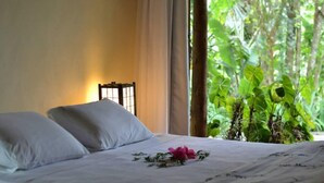 A bed with white linens and a pink flower, a nightstand with a lamp, and a view of lush greenery through a window. - Hotel Warabi (Resende)