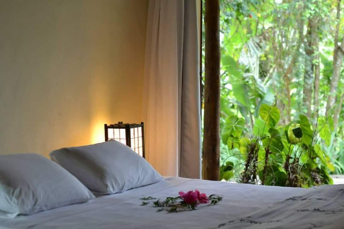 A bed with white linens and a pink flower, a nightstand with a lamp, and a view of lush greenery through a window.