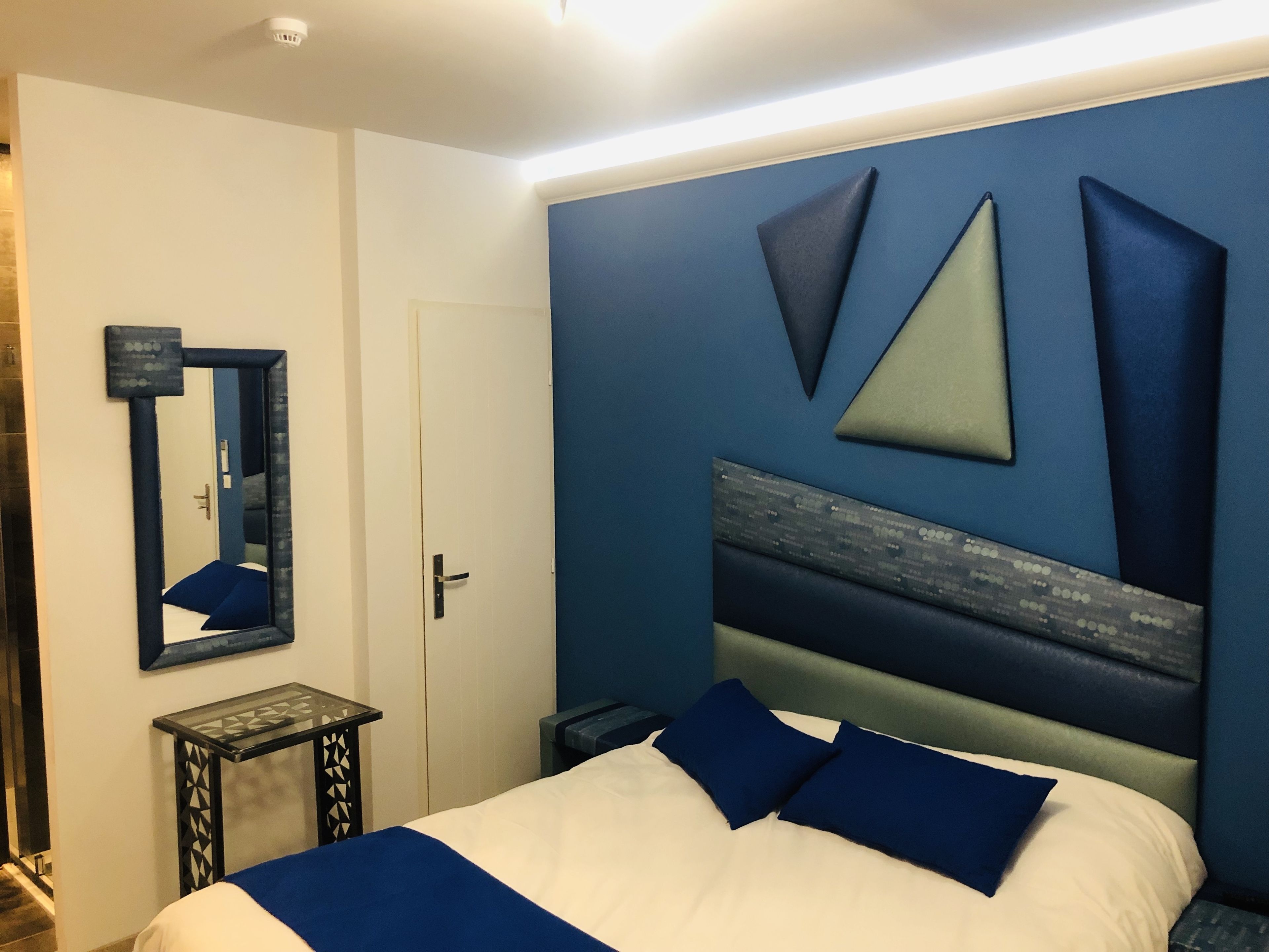 Quarto com cama, mesa de cabeceira, espelho e parede com padrões geométricos.