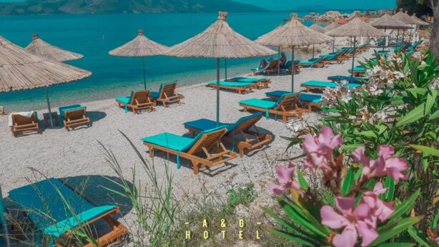 A beach with sun loungers and thatched umbrellas, a clear view of the sea, and a mountainous backdrop. - A&G Hotel (Orikum)