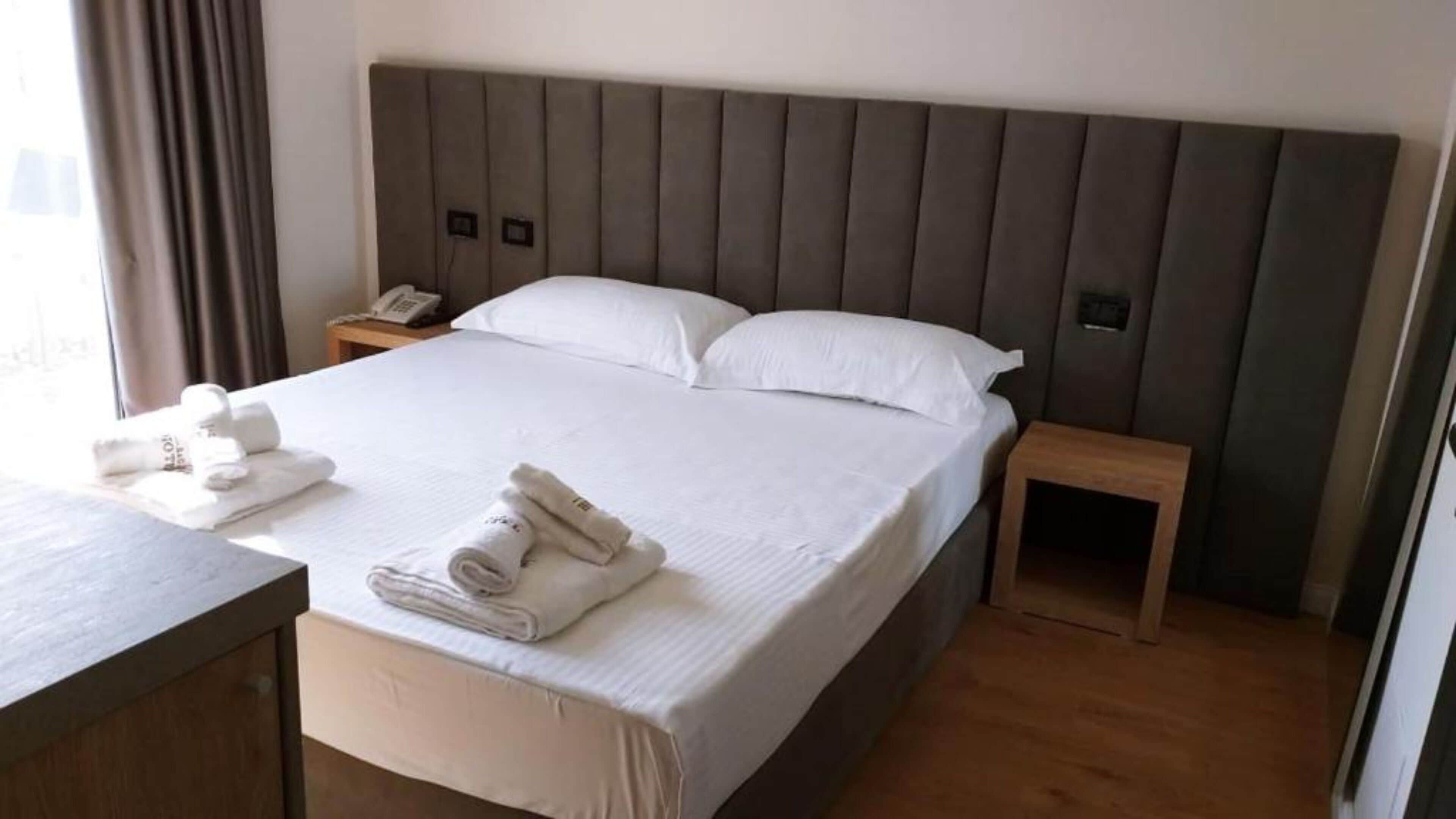 A neatly made bed with white linens and pillows, a wooden nightstand with a phone, and a wooden desk with folded towels.