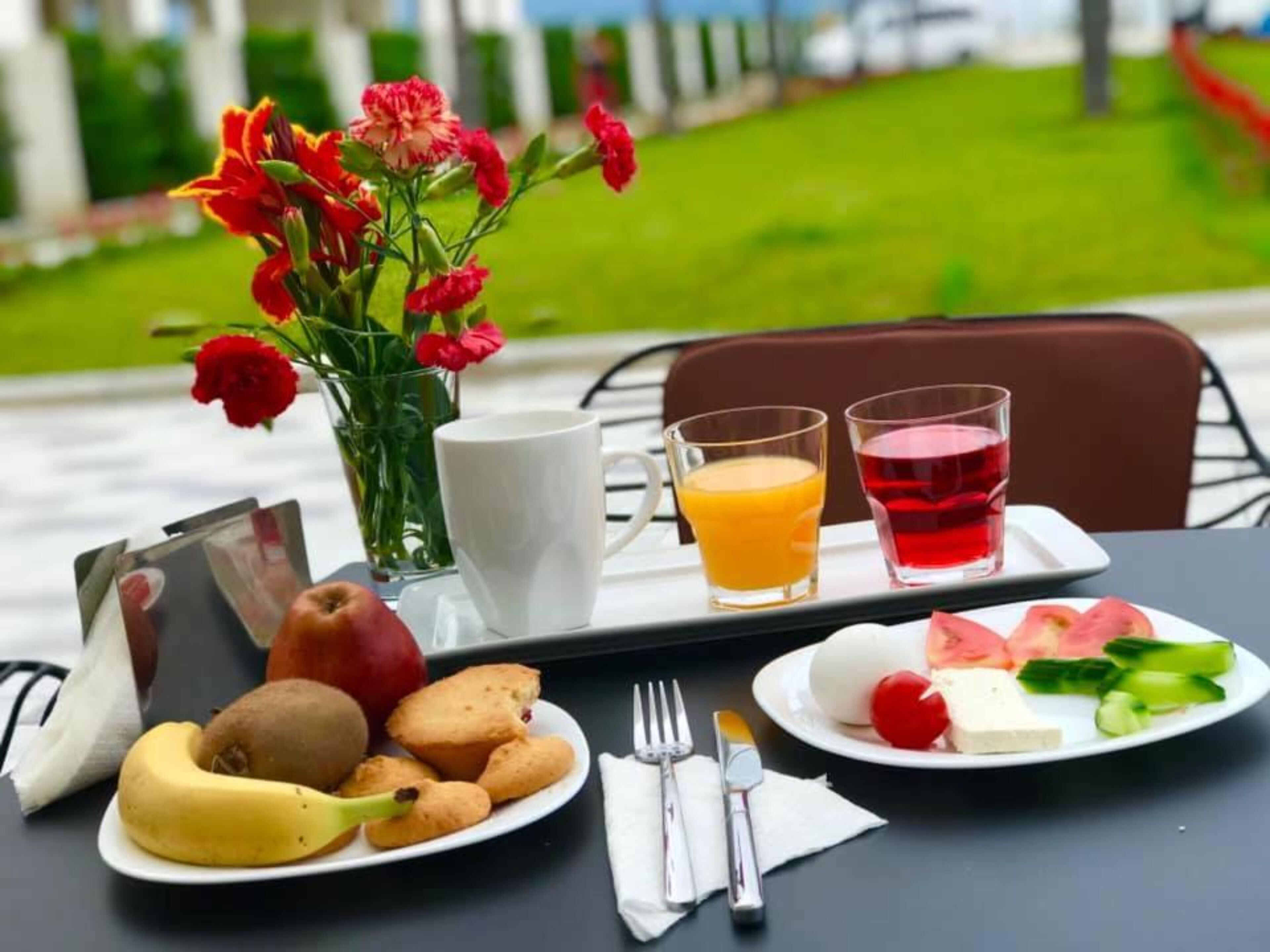 Desayuno con un plato de comida, un vaso de jugo de naranja, un vaso de bebida roja, un plátano, kiwi, galletas y un pequeño jarrón con flores.
