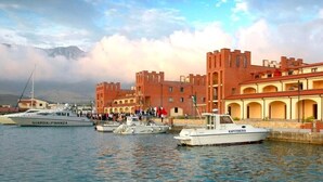A marina with boats docked, a large brick building with arched windows, and a mountainous backdrop. - A&G Hotel (Orikum)