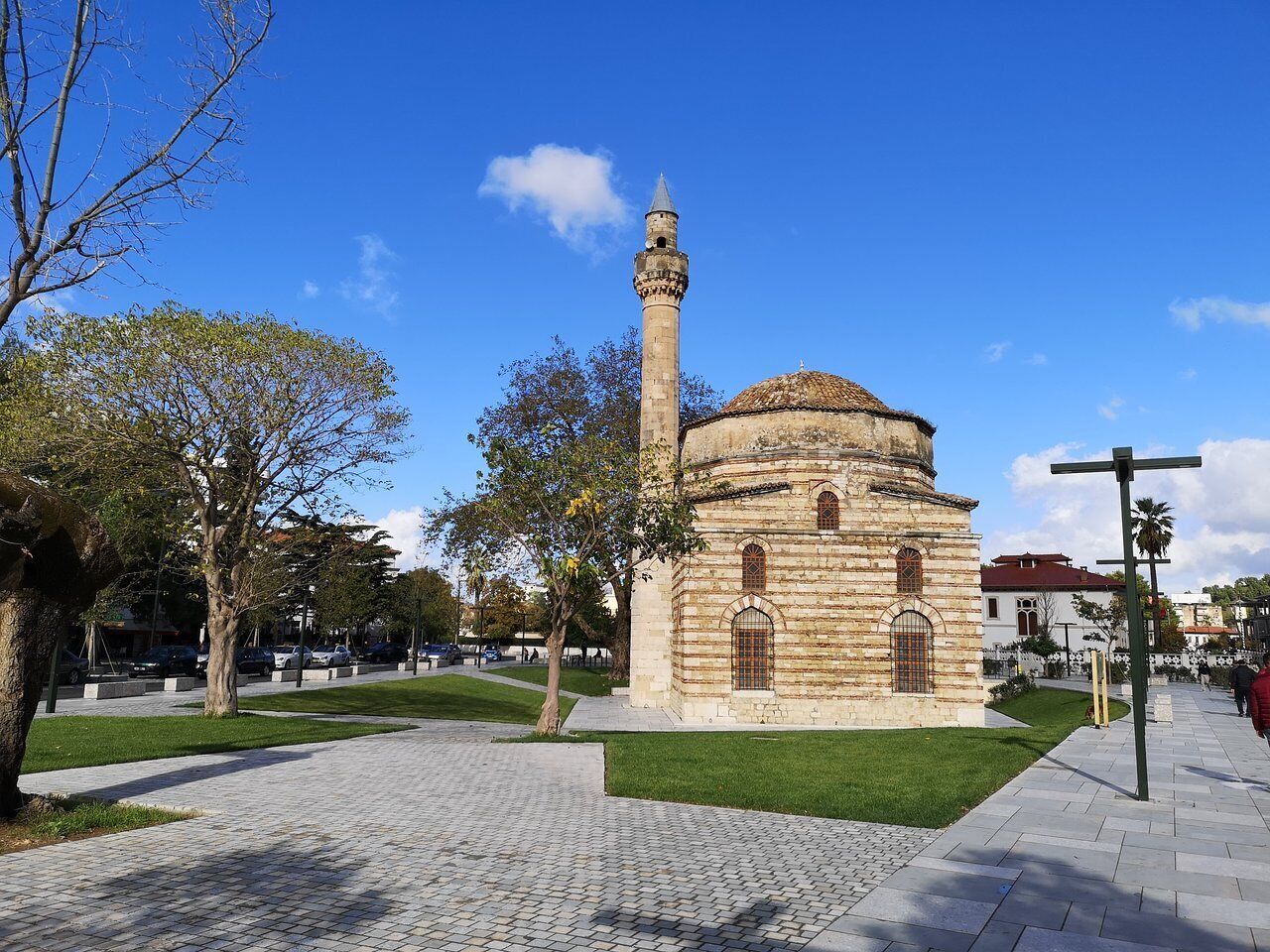 Ein historisches Gebäude mit einem markanten Minarett und einer Kuppel, umgeben von einer gepflasterten Fläche mit Bäumen und blauem Himmel.