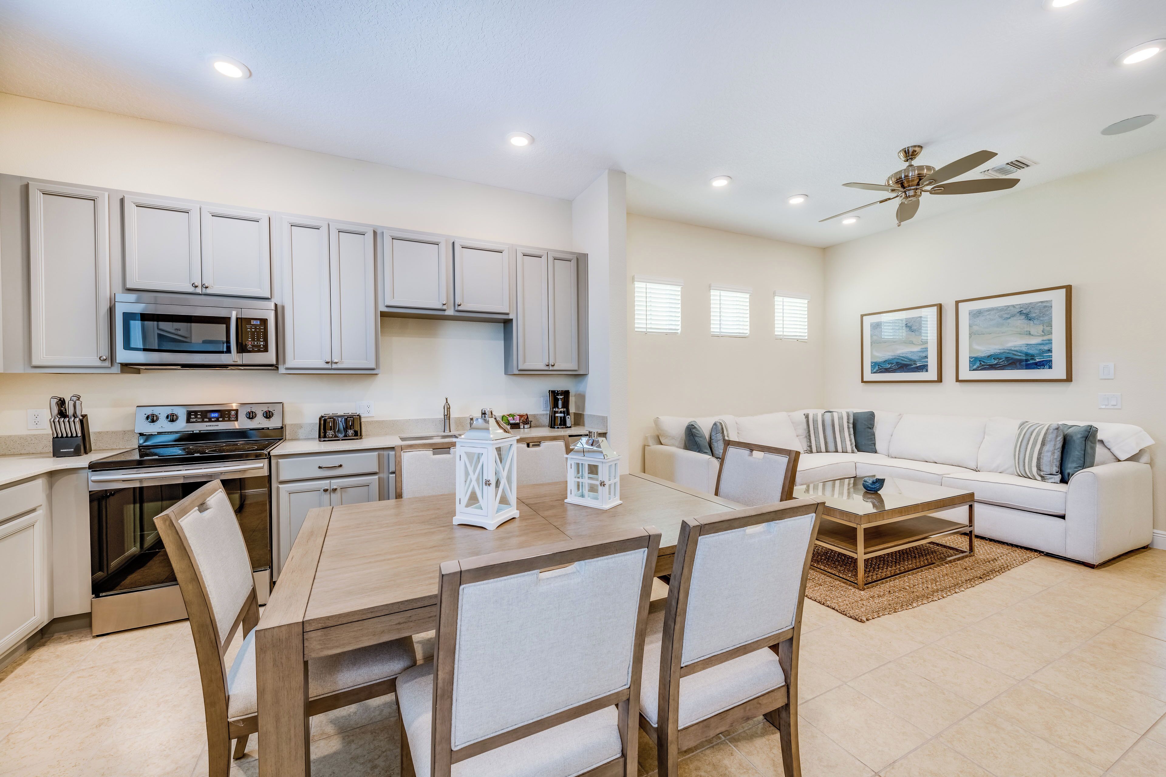 A modern kitchen with white cabinetry, a built-in microwave, and a stove. There is a dining area with a wooden table and chairs, and a living room with a white sofa and a ceiling fan.
