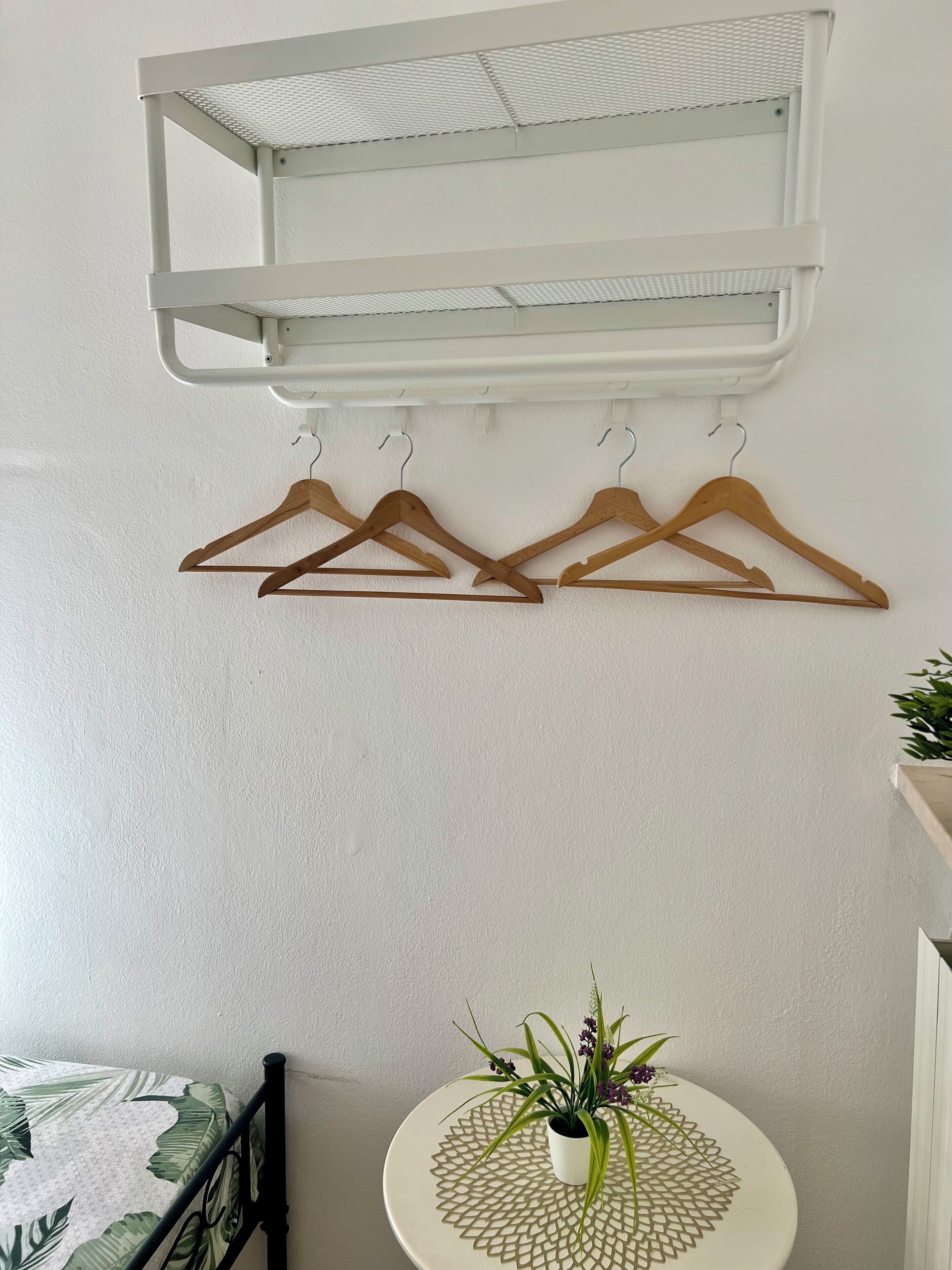 A white wall with a ceiling-mounted shelf and four wooden hangers. Below, a round table with a plant and a decorative mat is visible.