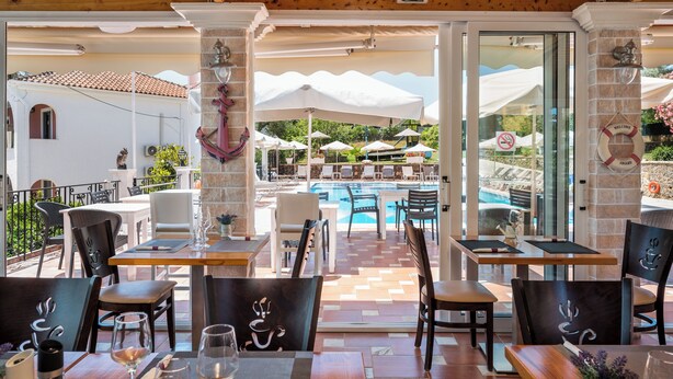 Un área de comedor al aire libre con mesas y sillas, una piscina y sombrillas al fondo.