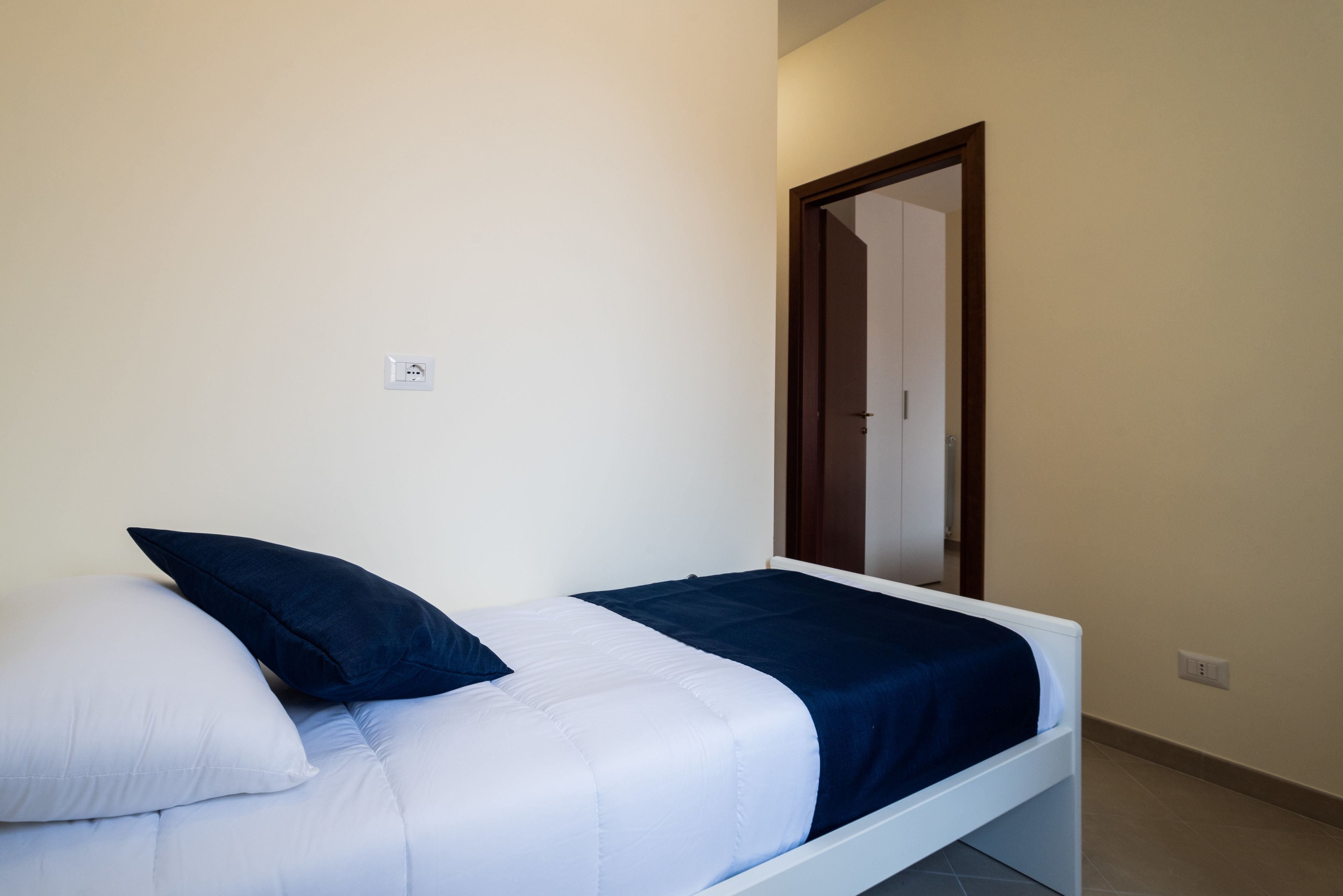 Una cama individual con ropa de cama blanca y azul oscuro, una pared blanca y una puerta de madera.