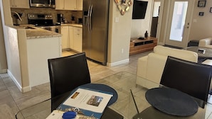 Eine moderne Küche mit Edelstahlgeräten, ein Essbereich mit Glastisch und ein Wohnzimmer mit Sofa und Fernseher.