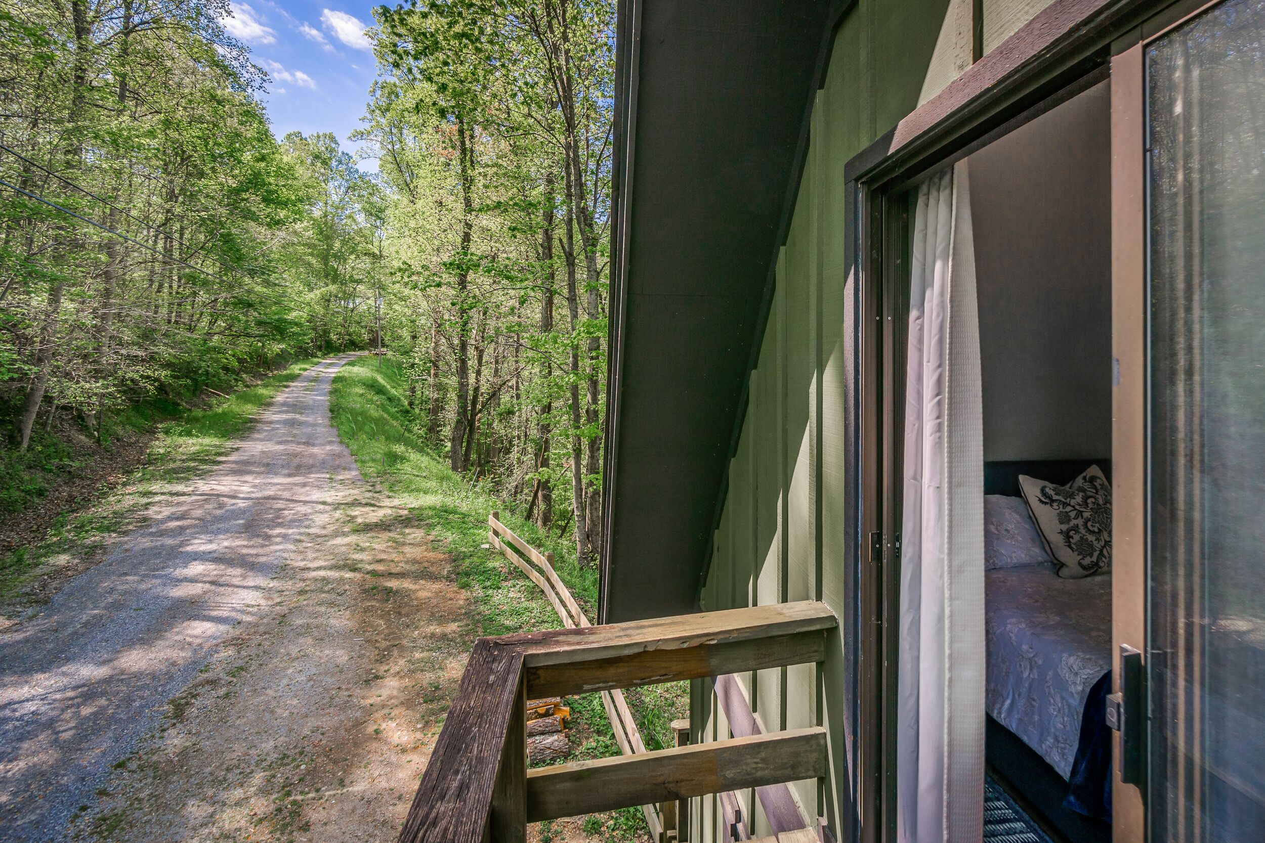 A wooden bench overlooking a forested path leading to a building with a bed visible through a glass door.