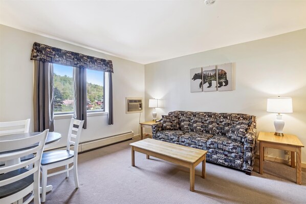 Ein Wohnzimmer mit einem gemusterten Sofa, einem Holztisch, einem weißen Sessel und einem Esstisch. Zwei Wandlampen und ein Fenster mit Vorhängen sind vorhanden.
