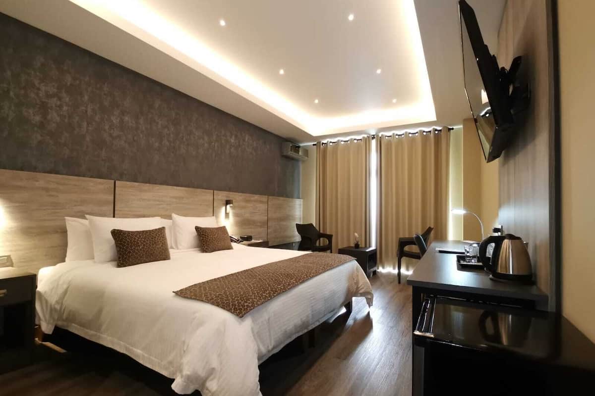 A modern hotel room with a large bed, bedside tables, a desk with a kettle, and a flat-screen TV mounted on the wall.