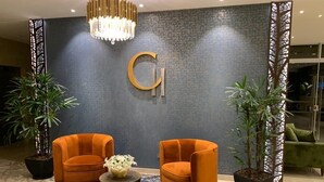 Un vestíbulo con dos sillones naranjas, una alfombra estampada, una mesa central con un jarrón y una gran 'G' en la pared.