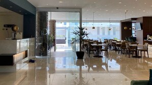 Un moderno vestíbulo de hotel con separador de vidrio, un mostrador de recepción y una zona de comedor con mesas y sillas.