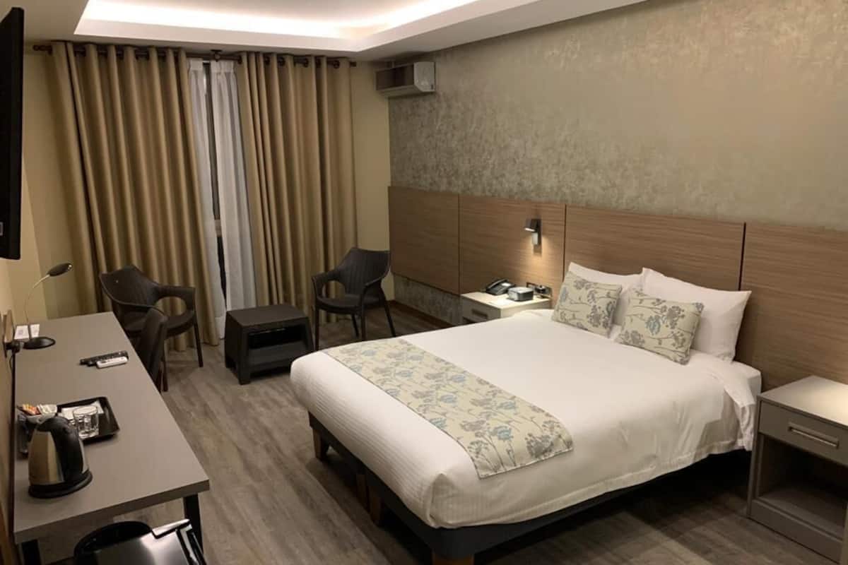 A hotel room with a large bed, a desk with a chair, a small table, and a nightstand with a lamp.