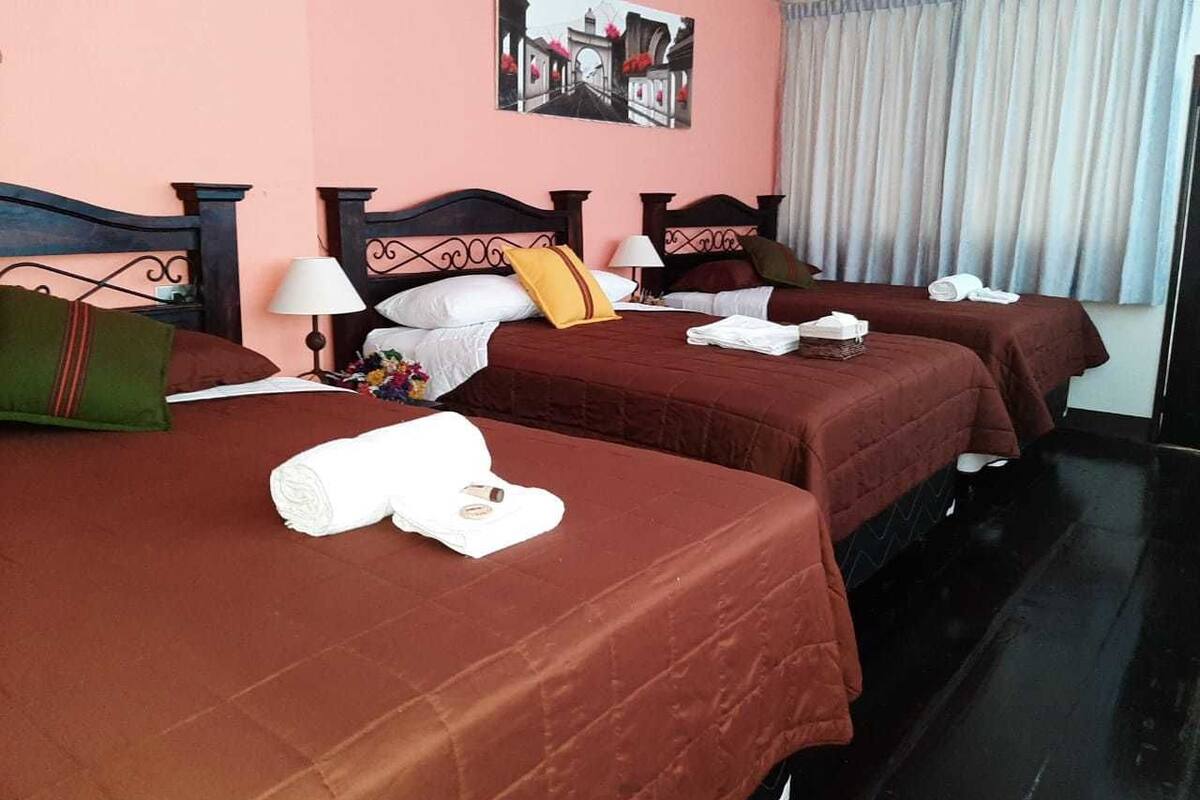 A hotel room with three beds, each with a red bedspread and white towels. There is a painting on the wall above the beds, and a window with curtains.