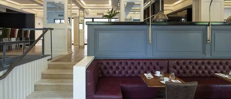 A modern interior with a wooden herringbone floor, a red leather booth, and a staircase with a glass railing.