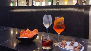 Una barra con una variedad de botellas, un tazón de papas fritas, un vaso de bebida roja y un vaso de bebida transparente.