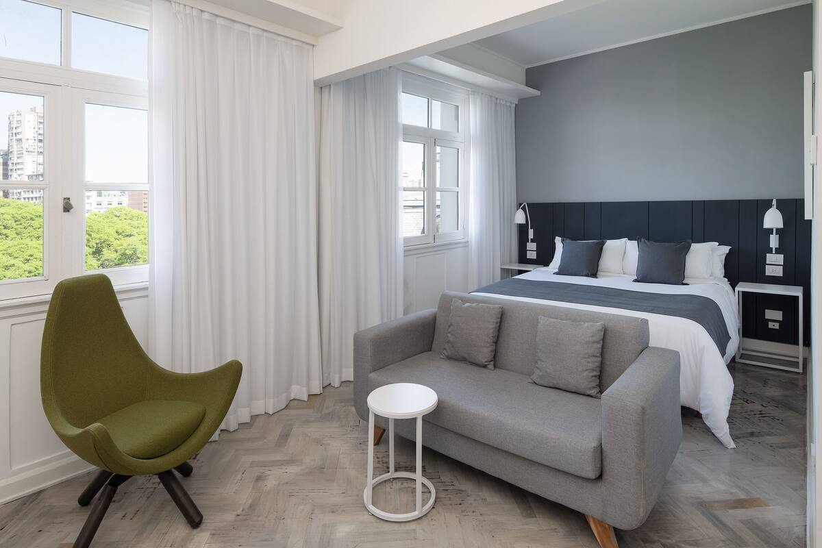 A modern hotel room with a bed, a grey sofa, a green chair, and a small white side table.
