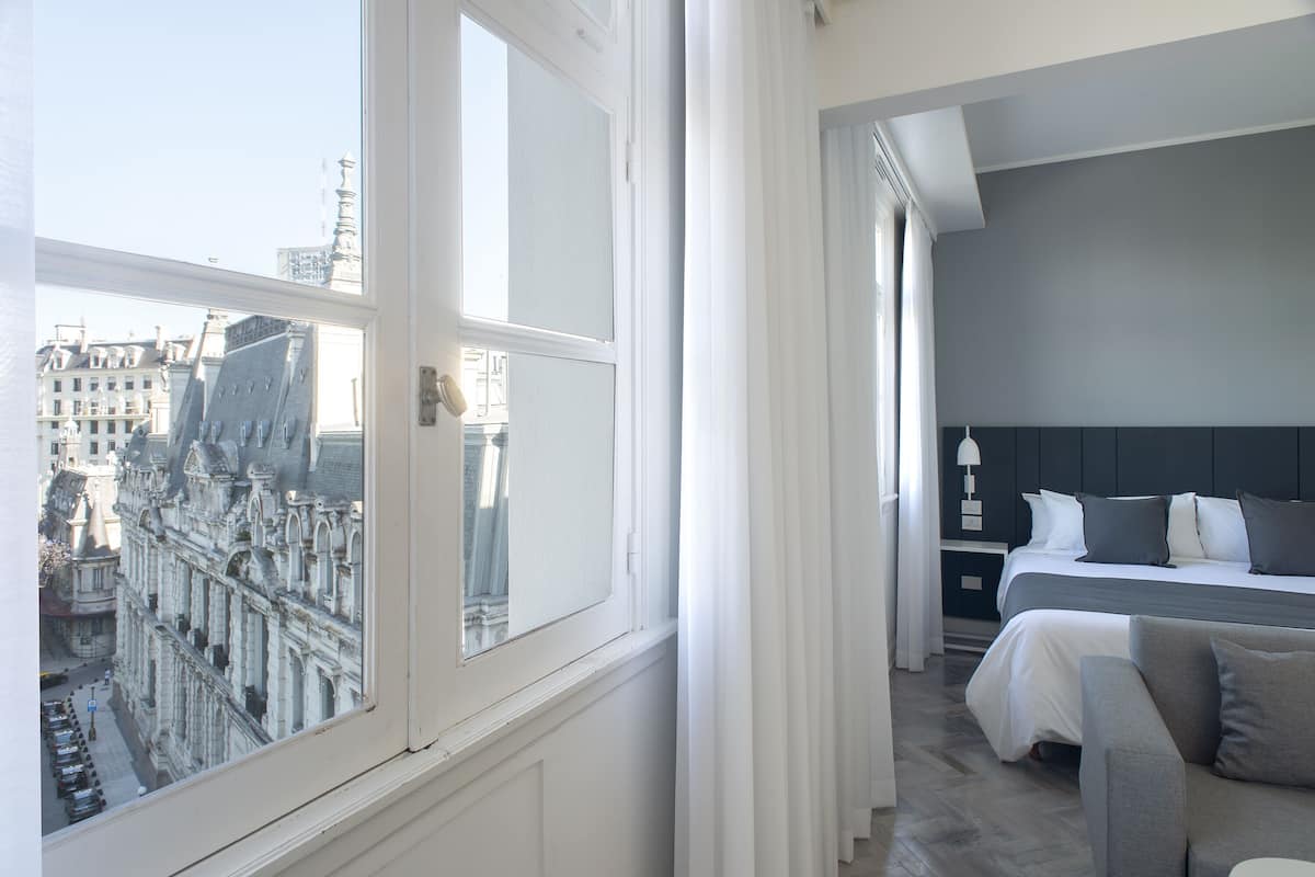 A hotel room with a large window, a bed with grey bedding, a bedside table, and a chair.