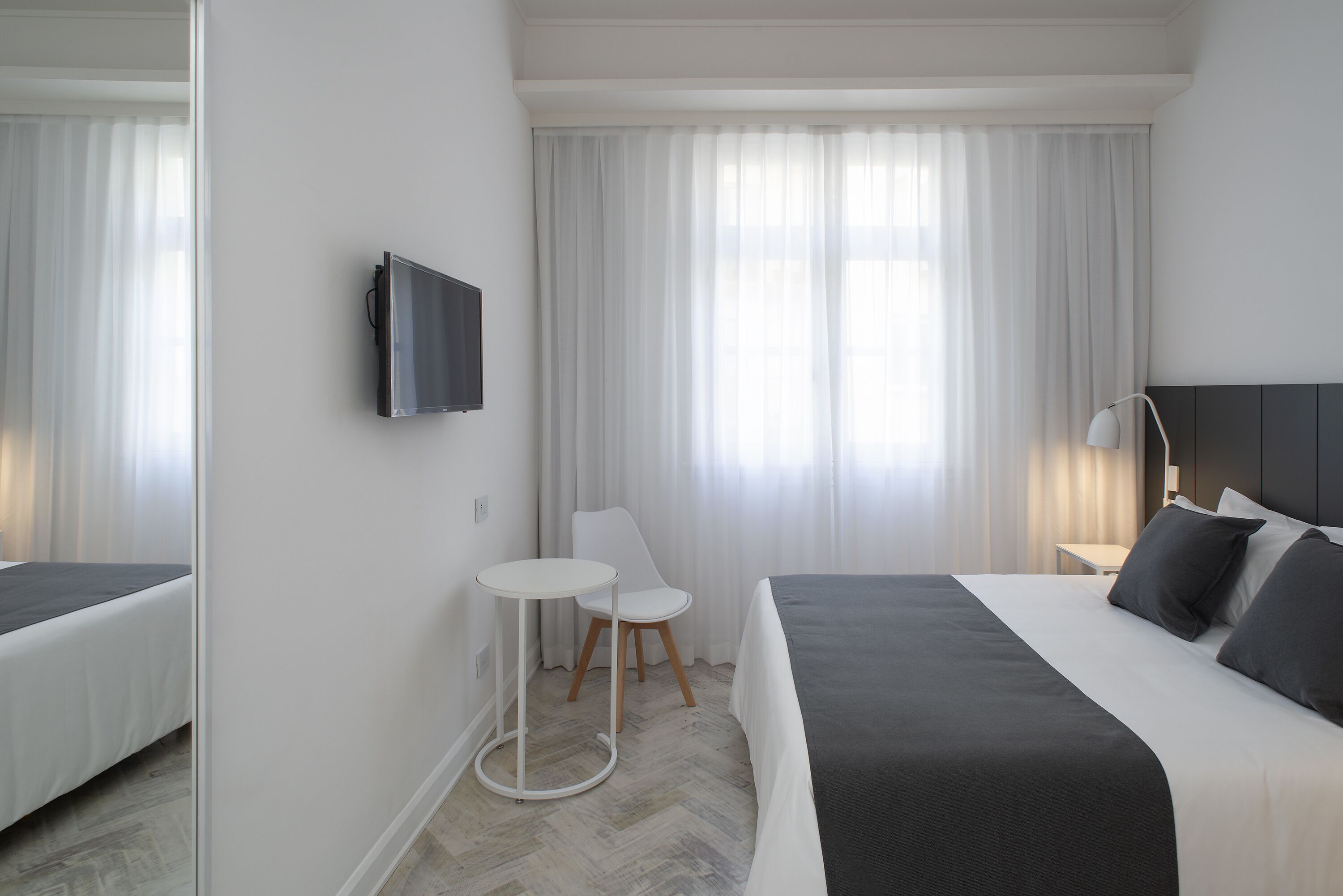 A modern hotel room with a bed, a small table, a chair, a TV, and a large window with curtains.