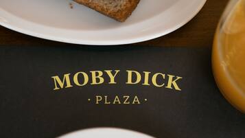 Una rebanada de pan integral sobre un plato blanco, un menú negro con 'MOBY DICK PLAZA' escrito en dorado, y un plato con manzanas en rodajas y un limón.