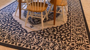 A dining area with a wooden table and four chairs, a patterned rug, and a doorway leading to a living room.
