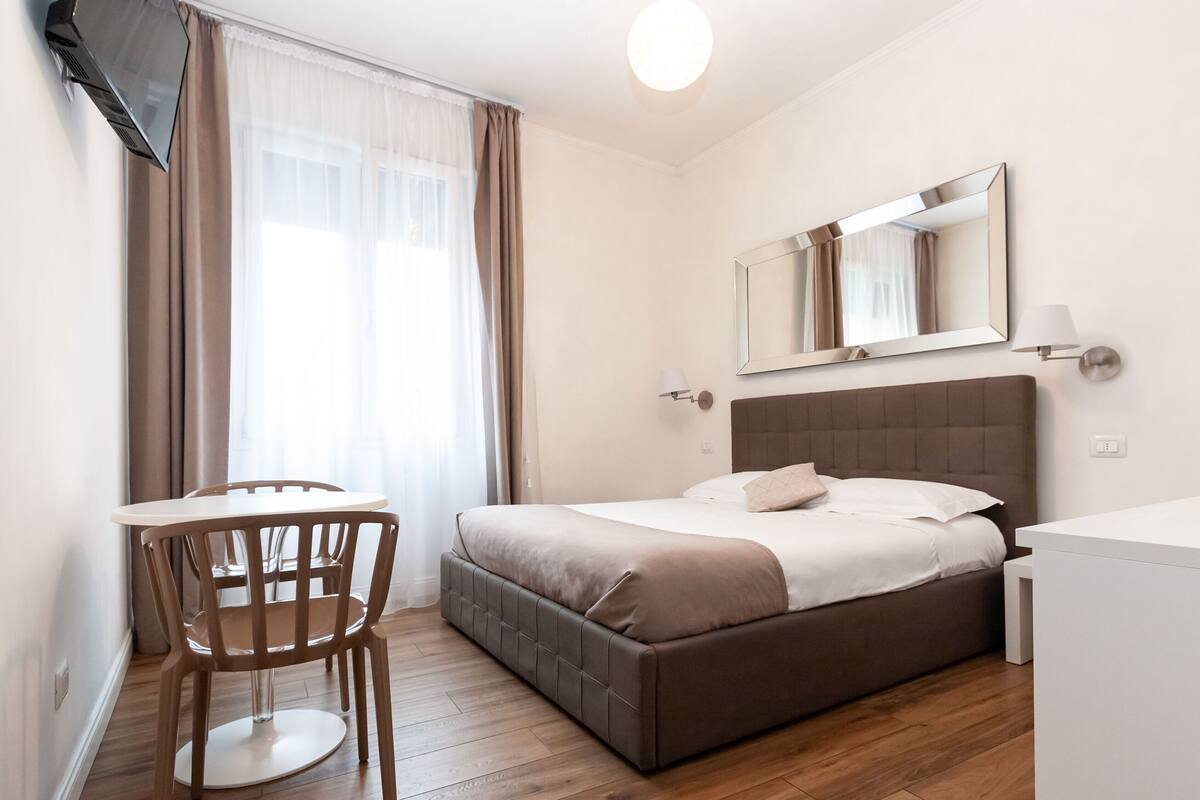 Een hotelkamer met een bed, een tafel met stoelen, een spiegel en een raam met gordijnen.
