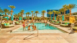 A pool area with lounge chairs and umbrellas, surrounded by palm trees and a modern building in the background.