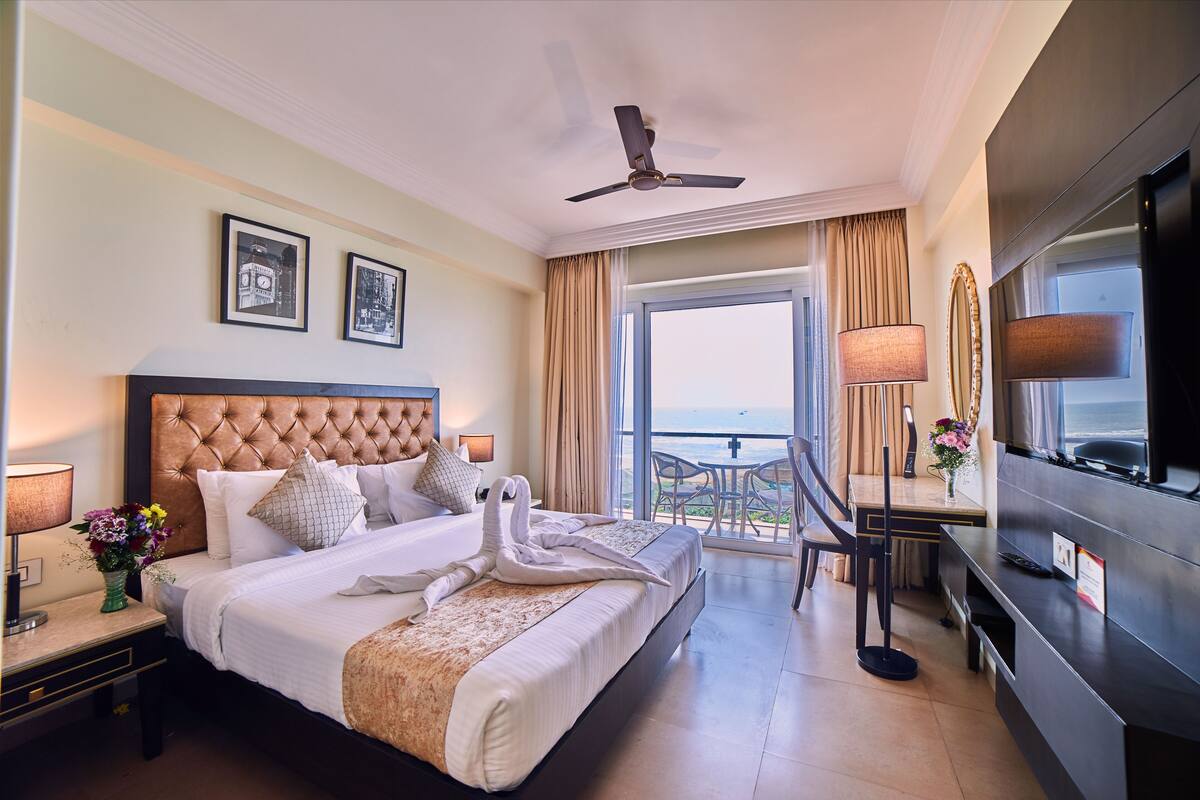 Habitación de hotel con una cama grande, televisión, escritorio con silla y balcón con vista al mar.