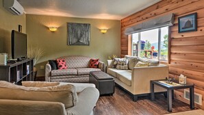 Ein gemütliches Wohnzimmer mit einer Couch, einem Hocker und einem kleinen Tisch. Ein Fernseher steht auf einem Lowboard, ein Fenster bietet einen Ausblick und eine Wandleuchte ist angebracht.