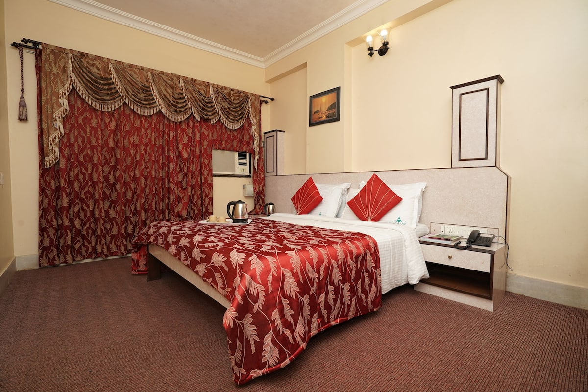 Quarto de hotel com cama, mesa de cabeceira, abajur e janela com cortinas.
