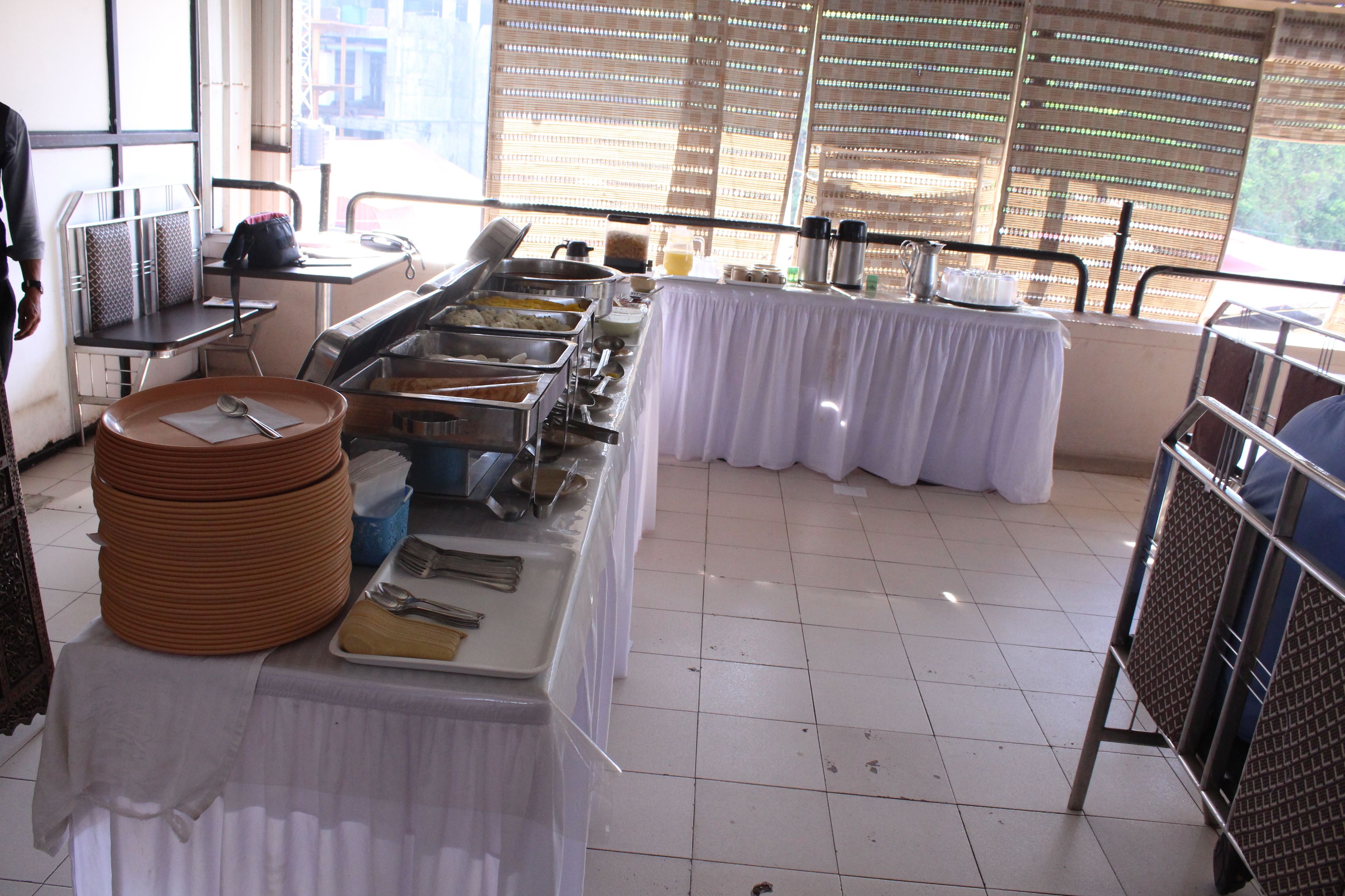 A buffet setup with multiple serving stations, a table with stacked plates, and a window with blinds in the background.