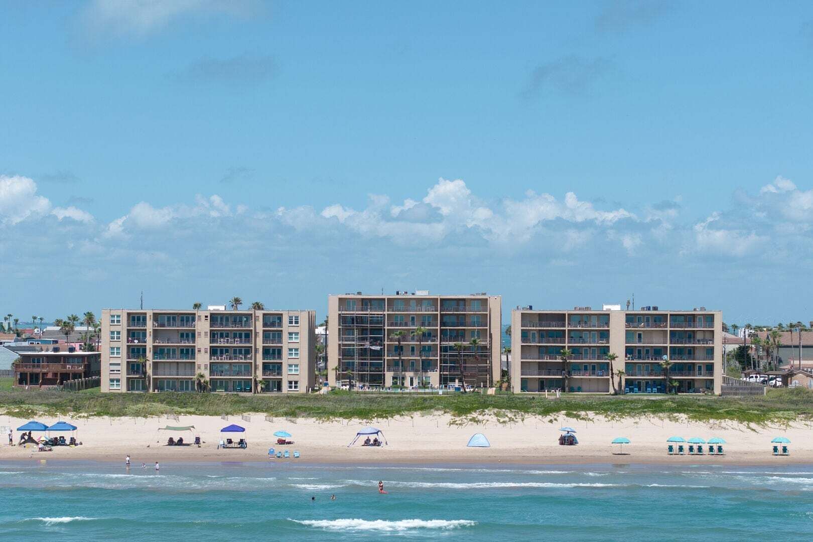 Um hotel à beira-mar com vários andares, uma praia arenosa em primeiro plano e um céu claro.