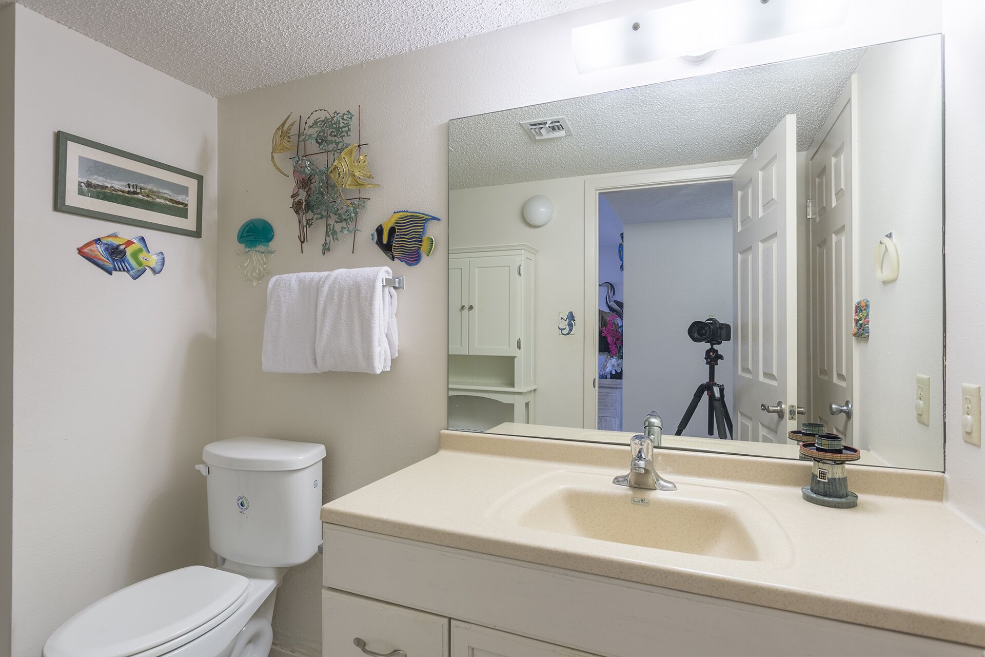 Ein Badezimmer mit Toilette, Waschbecken und Spiegel. Ein Handtuch hängt an der Wand und im Spiegelbild ist eine Kamera auf einem Stativ zu sehen.