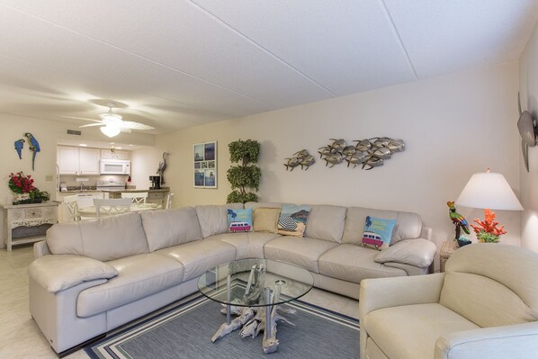 Een moderne woonkamer met een hoekbank, een glazen salontafel en een eethoek met witte kasten en een keuken op de achtergrond.