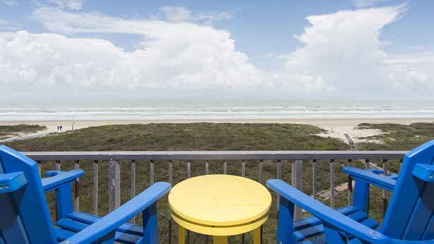 Una terraza con sillas azules y una mesa amarilla con vista a una playa que tiene una zona verde y un sendero que lleva a la orilla.