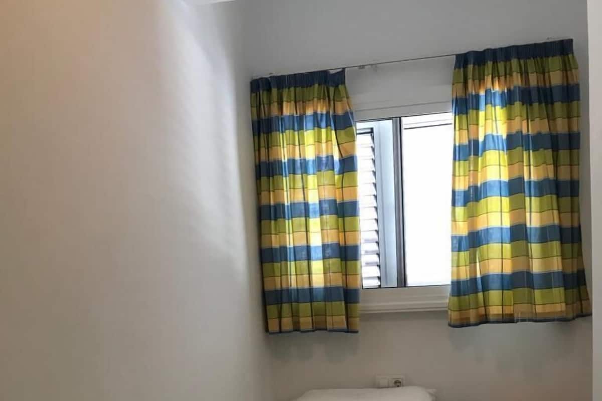 Una habitación pequeña con cama individual, ventana, cortinas a rayas y un banco.